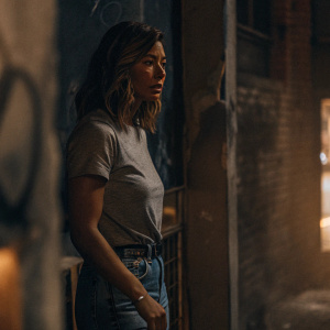 Jessica Biel, wearing a casual grey t-shirt and denim jeans, stands in a dimly lit alley. Graffiti-covered walls in the background.