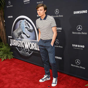 Nick Robinson, a young man with fair skin, at the premiere of 'Jurassic World'. Wearing casual striped shirt and sneakers. Dark backdrop with film logos.