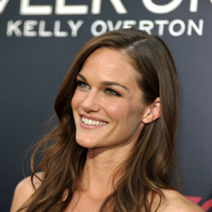 Kelly Overton, Caucasian actress, smiling warmly. Long brown hair cascades down her shoulders. Dark background with bold white text. Close-up shot highlighting her serene expression.