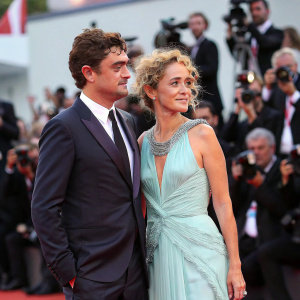 Riccardo Scamarcio in a dark suit and Valeria Golino in a light blue gown at the Venice Film Festival. Crowds and paparazzi surround them.