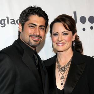 Danny Nucci and Paula Marshall at a Hollywood gala. Danny, with dark hair and beard, dressed in sleek black suit. Paula, elegant in a black blazer and statement necklace. Both smile against a plain white backdrop, surrounded by GLAAD banners.