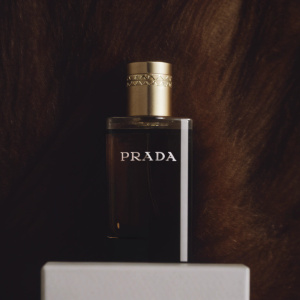 Photograph showcasing the Prada L'Eau Ambree perfume bottle. The bottle is sleek and elegant, with a black body and a gold-toned cap. Laid out on a pristine white packaging box, which displays the brand name 'PRADA' in bold font.
