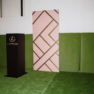 Promotional stand for Sabror de Lujo gala. Modern design with sleek black and gold accents. Geometric patterned backdrop, dark and textured wall. Lexus logo prominently displayed.