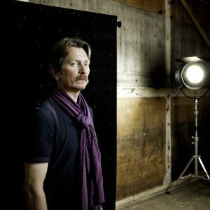 Middle-aged actor Gary Oldman, with a serious expression and neatly trimmed mustache. Wearing a dark purple scarf. Standing against a textured black backdrop, spotlighted face and upper body. Moody and dramatic lighting.