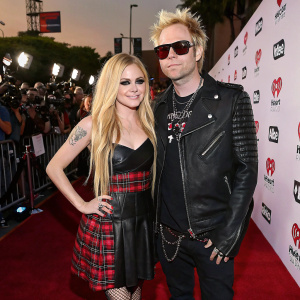 Avril Lavigne and Mod Sun at the iHeartRadio Music Awards, red carpet scene. Lavigne in a sleek black dress with plaid detailing, Mod Sun in a stylish black leather jacket and dark shades.