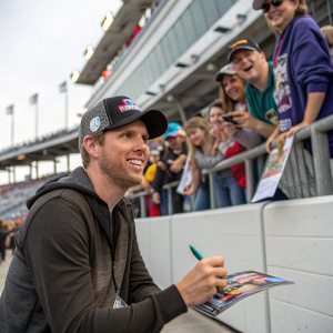 Denny Hamlin, in casual wear, posing with autograph in hand at fan meet and greet. Background: grandstand, fans cheering