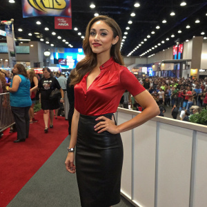 Lindsey Morgan at the Comic-Con event, dressed in a sleek red blouse and black skirt. The backdrop is a bustling convention hall filled with fans and cosplayers.