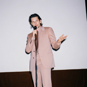 Bradley Cooper in a formal suit, dark hair, blue eyes. Press conference with stark white backdrop.