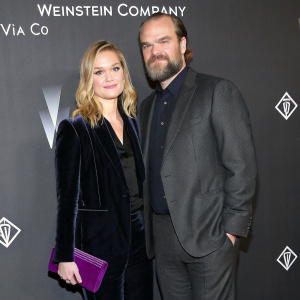 Julia Stiles and David Harbour at a glitzy Hollywood gala. They stand out in a sea of stars. Julia in a black velvet blazer paired with a dark top and purple clutch. David is in a sharp dark gray suit. The backdrop is adorned with The Weinstein Company and VIA CO logos. 