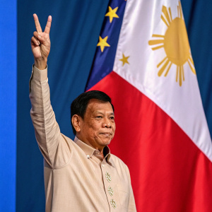 Rodrigo Duterte, ex-President of the Philippines, giving a peace sign with a serious look. Blue and red backdrop with the Philippine flag
