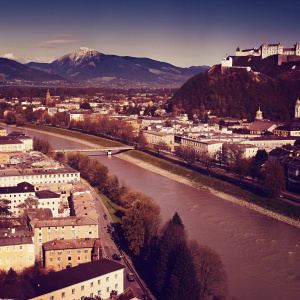 From a bird's eye view, Salzburg, Austria twinkles with the Danube river weaving through it. Hohensalzburg Castle stands prominently, amidst a blend of residential homes and bustling commercial districts, interspersed with lush green parks. Behind lies a panorama of majestic snow-capped mountains, against a backdrop of a radiant blue sky.