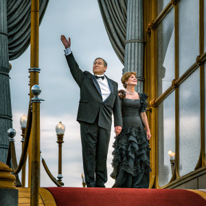John Goodman and Annabeth Hartzog attending the Met Gala. John in black tuxedo, Annabeth in elegant black gown with ruffles, standing on glamorous red carpet.