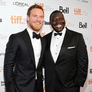 Michael Fassbender and Steve McQueen, dressed in black tuxedos, at a gala event. Smiling broadly, against a minimalist white backdrop with sponsor logos. Toronto International Film Festival.