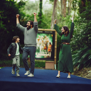 The Lopez family, Mario, Dominic, and Gia Francesca, posing joyfully at the Jumanji premiere. Mario in gray sweater, Dominic in gray shirt, Gia in green dress with belt. Blue carpet with movie poster backdrop.