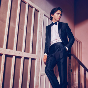 Timothée Chalamet, dressed in a black tuxedo, with slicked-back hair, standing alone in a dark alley. No background, just a silhouette against black