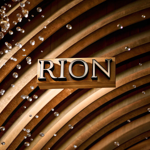 Sleek 'Rion' logo made of reflective material with large, curved segments. Set against a dynamic backdrop of tiny, translucent water droplets in motion.