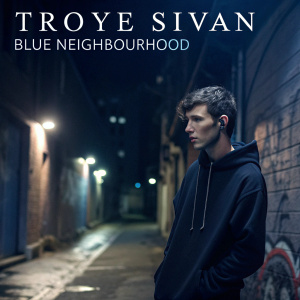 Album art for Troye Sivan's 'Blue Neighbourhood'. The singer is seen in a gritty urban setting, wearing a black hoodie. Dark alleyway with graffiti backdrop