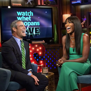 Andy Cohen and Naomi Campbell laughing heartily. Andy in a dark suit and tie, Naomi in a vibrant green dress. They are seated on stylish chairs with props and a large screen in the background. Watch What Happens Live set