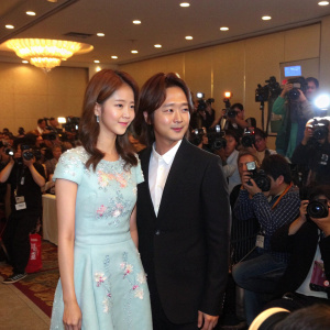 Yoona and Jang Geun-Suk at the Love Rain press conference. She's wearing a light blue dress with floral details. He's in a black suit. Crowded room with flashing cameras