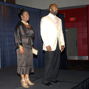 Kalilah Allen-Harris and Jonathan Slocumb at the 39th NAACP Image Awards. Kalilah in brown dress and jewelry, Jonathan in white suit with pink-patterned tie. Stage with red and gold backdrop