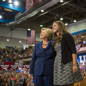 Hillary Clinton, in a dark blue suit, stands with a woman in a patterned skirt at a crowded political rally. Various attendees with diverse backgrounds gather inside the hall.