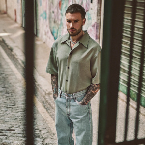 Close-up candid photo of Liam Payne. Wearing a green shirt and denim jeans. Liam has short dark hair and a stubble beard. Visible tattoos on his arms. He is standing in front of a street with colorful graffiti walls.
