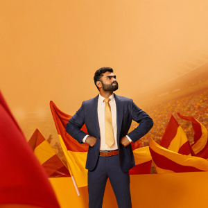 Photograph of Virat Kohli, cricket star, wearing a navy suit, gold tie. Crowded stadium background, vibrant with fans and flags.