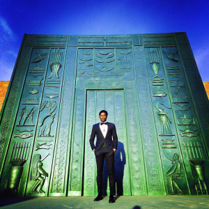 Chadwick Boseman at the premiere of Gods of Egypt. He is dressed in a tuxedo, standing in front of an elaborate, ornate backdrop depicting ancient Egyptian motifs. Black and white photo.
