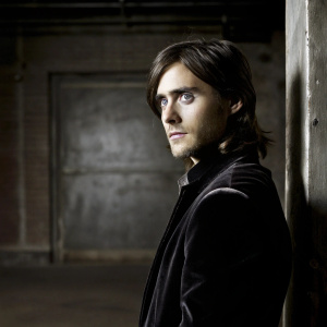 Renowned actor Jared Leto standing solemnly. Dark hair cascading, intense blue eyes. Dressed in a black velvet jacket, standing in front of an all-black backdrop. Gently lit by a soft glow.