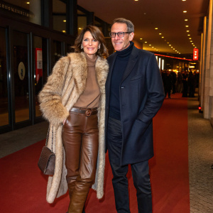 Gerit Kling and Wolfram Becker at Berlin Film Festival premiere. Gerit in fur coat, brown leather pants, and boots. Wolfram in dark navy blazer and turtleneck. Warm, ambient lighting and structured architecture. Candid style.