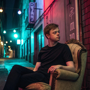 Dane DeHaan, dressed casually in a black t-shirt, sitting on a vintage armchair. Neon-lit urban alleyway in the background.
