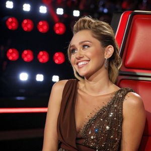 Miley Cyrus, host of The Voice, smiling brightly. Her brown dress is adorned with sparkling elements. Behind her, a sleek red chair stands out against the bright studio lights.