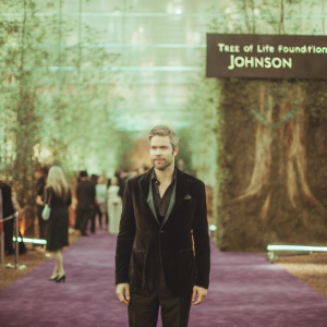 Ryan Serhant at a fundraising gala. Wearing a sleek black suit with a dark jacket. Standing in front of a vibrant green backdrop with various sponsor logos including Tree of Life Foundation and Johnson. The venue features a royal purple carpet and an array of guests.