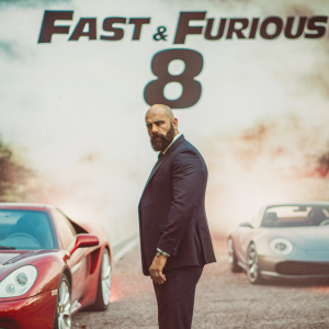 Dwayne Johnson in character, standing in front of 'Fast & Furious 8' movie poster. Serious expression, wearing a dark suit. Bearded, against a backdrop of sleek cars and co-stars.