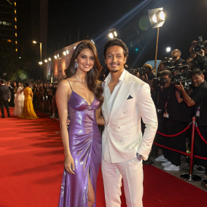 Tara Sutaria in a shimmering purple dress and Tiger Shroff in a sharp white suit at the Filmfare Awards ceremony. They are standing on a red carpet surrounded by flashing cameras and excited fans.