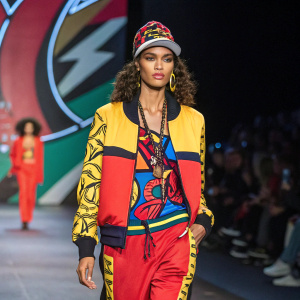 Hispanic model strutting on New York Fashion Week runway. Wearing Rocawear-inspired outfit, vibrant backdrop.