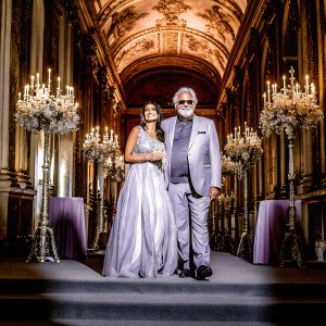 Elisabetta Gregoraci and Flavio Briatore at an elegant charity gala. The venue is a grand hall adorned with chandeliers and floral arrangements. Elisabetta exudes warmth in a flowing evening gown, while Flavio sports a stylish suit and displays a welcoming smile.
