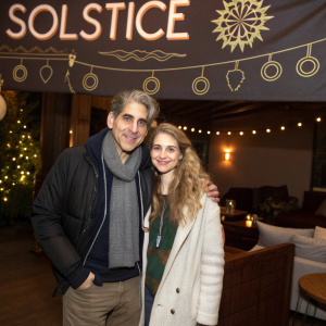 Michael Imperioli and Eden Wexler at a cozy indoor event. Wearing casual outfits. Standing in front of a 'Solstice' themed banner.