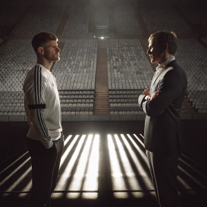 Timo Werner and Thomas Tuchel standing on a bright stadium floor. A sea of seats in the background. Evening lights casting shadows.