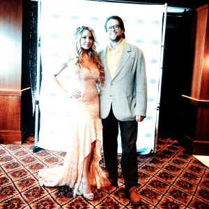Ayleen Mujica and Mauro Menendez attend an award ceremony. Ayleen in a dazzling floor-length gown with ruffled details. Mauro in a gray blazer and yellow shirt. They pose together on a patterned carpet in front of a backdrop with event logos and text.