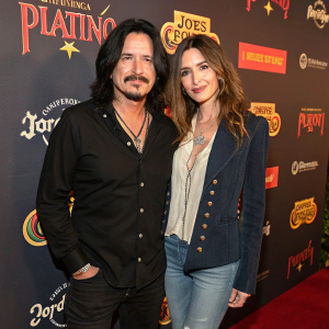 Gilby Clarke and Daniella Clarke at Jose Cuervo Platino event. Both dressed in casual attire, Gilby in a black shirt, Daniella in a buttoned blouse and jacket. The backdrop features vibrant branding and logos.