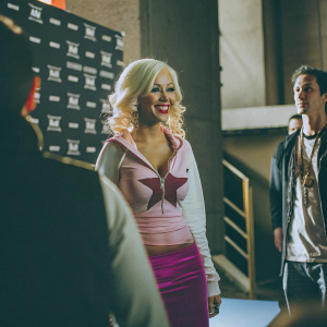 Christina Aguilera, blonde, in a casual outfit, smiling. Pink star design top, magenta skirt. Event backdrop with branded logos.