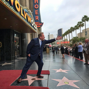 John Goodman, in a dark suit, playfully standing on a star on the Hollywood Walk of Fame. The iconic Hollywood sign and bustling crowd in the background.