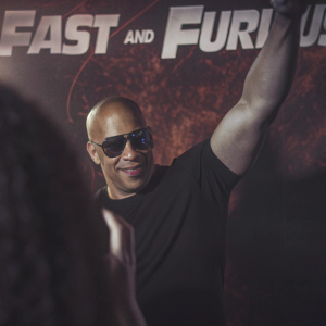 Vin Diesel, bald head, dressed in black t-shirt and dark sunglasses, posing at the Fast and Furious premier. Vibrant backdrop with fast and furious logo