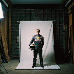 Sebastian Vettel, dressed in a full race suit, standing in front of a white backdrop with the Red Bull logo.