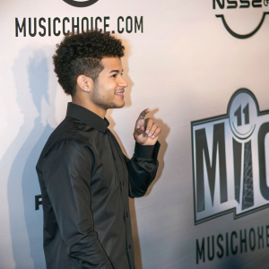 Jordan Fisher, youthful Hispanic male with curly hair, standing at a music event. Wearing a black shirt, relaxed pose. The backdrop features bold logos and text for MusicChoice.com.