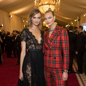 Cara Delevingne and Karlie Kloss at a high-profile fashion gala. Both in stunning evening wear, Cara in a black lace dress, Karlie in a plaid suit. The venue is luxurious, with a sea of attendees dressed in the latest trends.
