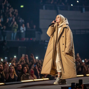 Billie Eilish on stage at the Brit Awards. Wearing oversized beige coat. Crowd cheers in the background