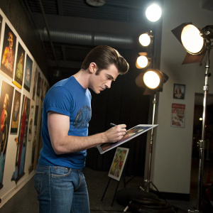 Drake Bell, dressed in a blue t-shirt and jeans, signing autographs. Bright studio lights, posters of his shows all around.