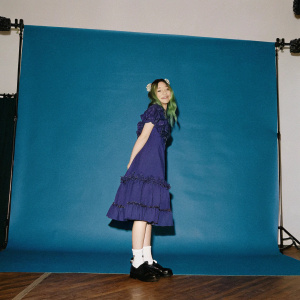 Lola, the character from Kiko Run, with green hair and a friendly smile. Purple dress with ruffled skirt. Standing on a white stage with bright spotlights.